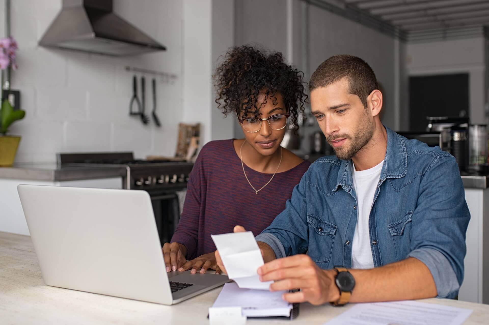 A man and woman sitting at a kitchen table with a laptop, looking at a receipt and engaging in a discussion.