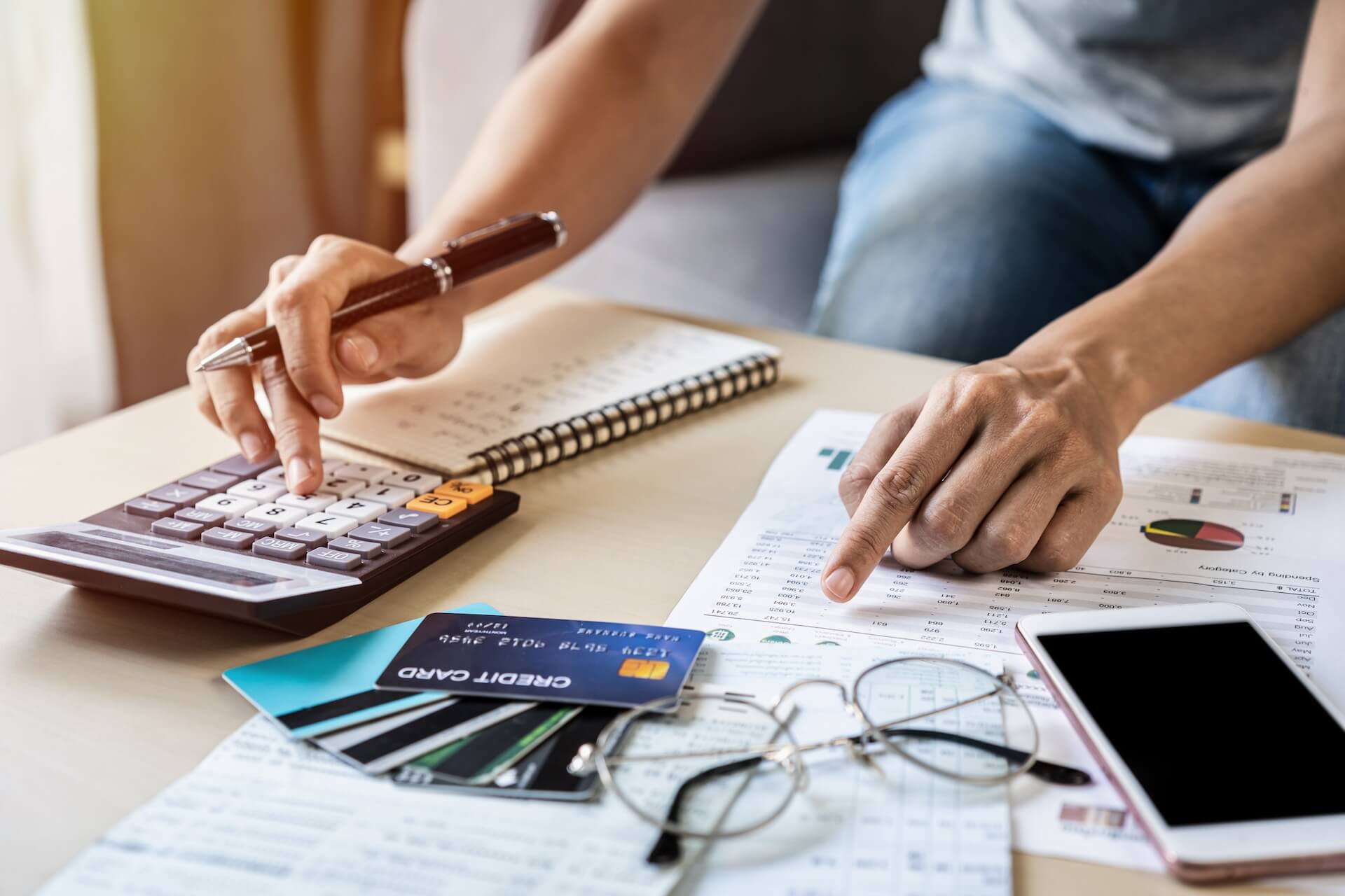 A person sitting at a desk using a calculator and holding a pen with a document, notepad, and credit card next to them.