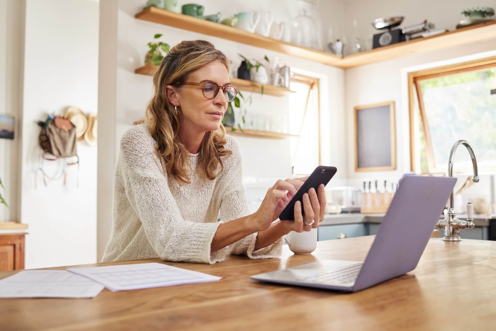 A woman with glasses sits at a kitchen table, using her phone with a laptop, documents, and a coffee mug next to her.